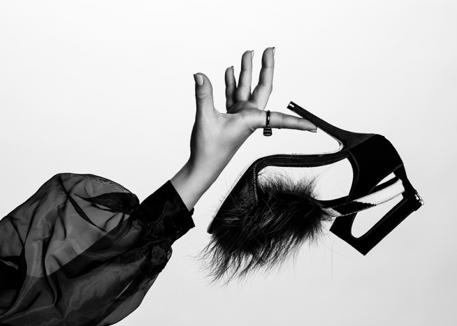  – Černobílá fotografie boty na podpatku s chlupatou špičkou, kterou drží žena v průsvitné volné halence